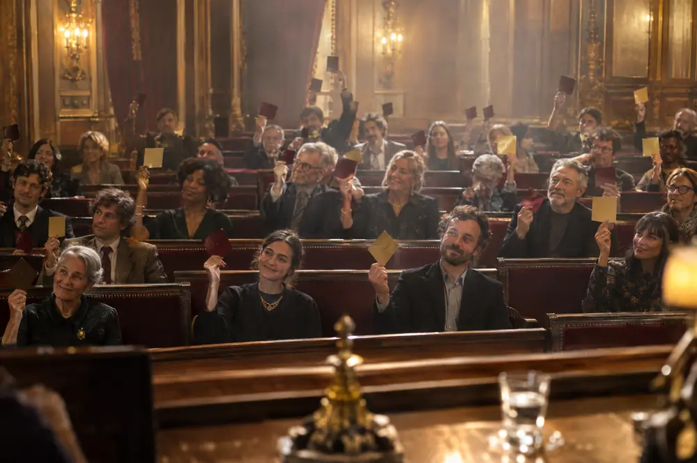 Le public d’une audience lève des cartons bordeaux et dorés dans un prétoire aux boiseries dorées